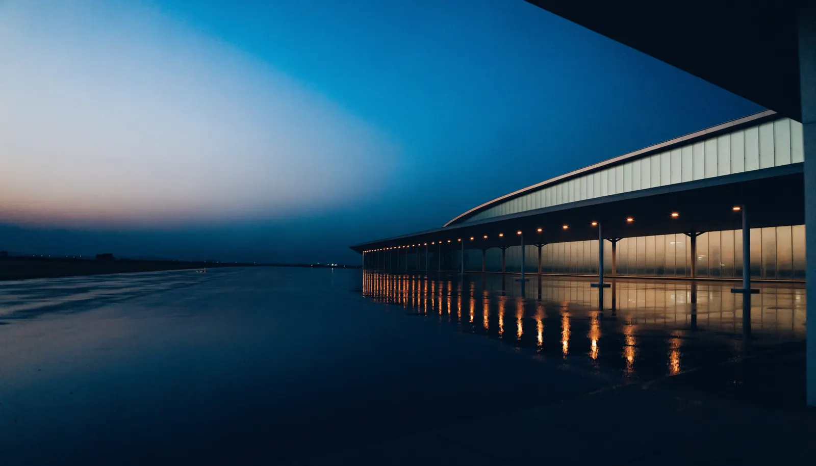 Terminal d'aéroport au crépuscule, façade vitrée reflétée sur le parvis mouillé